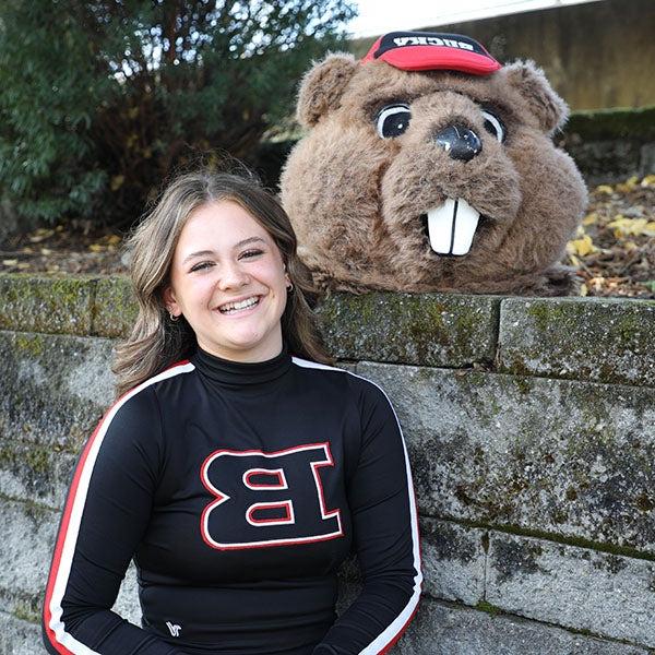 Nina穿着她的啦啦队运动衫，在巴拉德高中的吉祥物服装旁边微笑着拍照 
